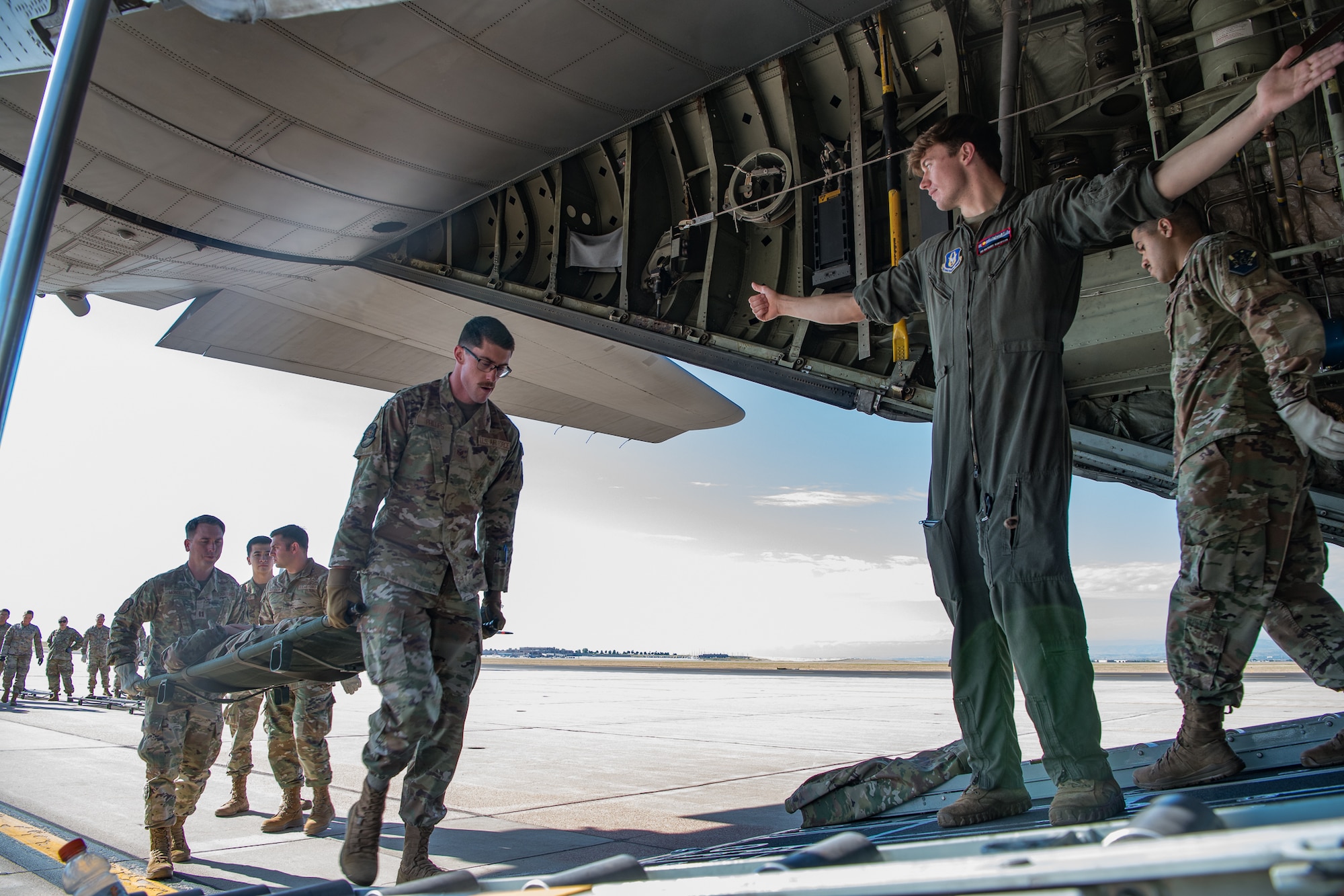 An Airman in a flight suit extends his arm toward an aircraft cargo bay to direct two Airman carrying a patient on a litter.