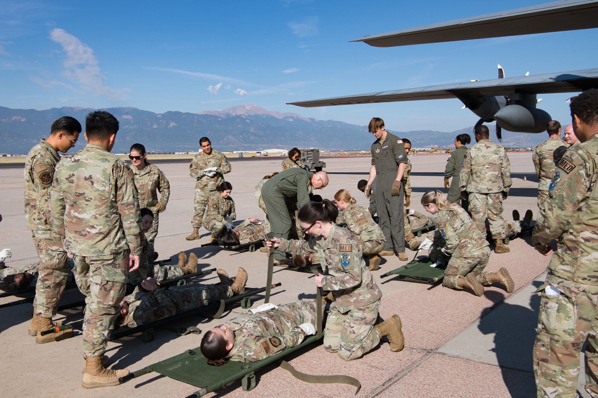 A group of Airmen securing their peers into litters on an aircraft parking ramp with mountains in the background and a C-130 wing behind them.