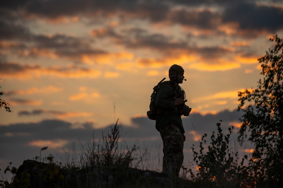 A soldier wearing gear and a headset stands at the top of the hill during twilight.