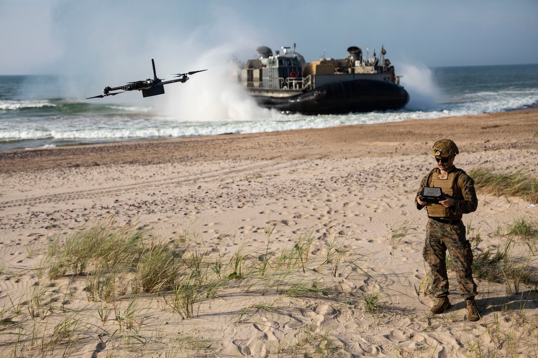 A solider pilots an unmanned aerial system on the beach while an amphibious landing craft comes ashore behind him.