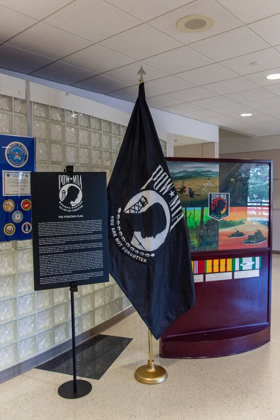 A black poster on a tall pole with white letters, the POE/MIA logo is next to a black flag with the POW/MIA logo which is next to a four panel painting of soldiers at war with campaign badges beneath it in a large foyer up against a white glass bottle wall.