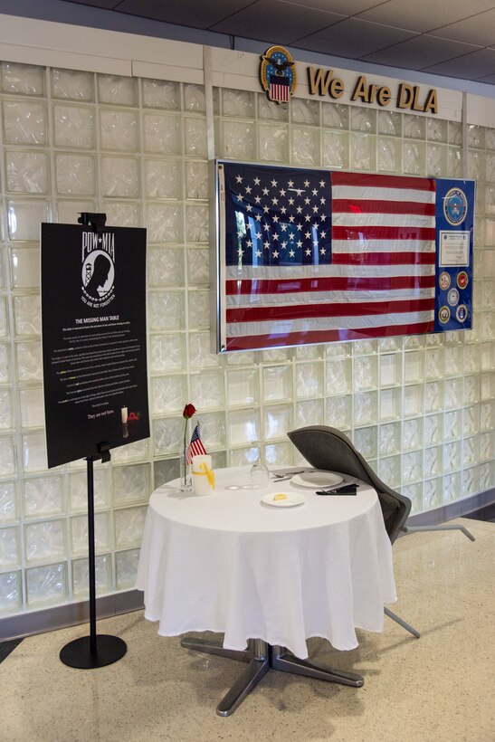 A table with a white tablecloth and a black poster sits next to a white glass bottle wall in a large foyer. The table is empty but there is a rose, a lemon on a plate, a table setting and a salt shaker on it. The black poster is held on a tall pole.