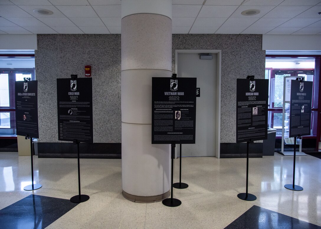 A display of posters with text white on black tells the stories of POW/MIA Day in a large vestibule area with doors to the outside on one side and a glass bottle wall on the other with an American Flag with the words We Are DLA above it. There is a table with a white cloth with a plate and other object on it. The table is empty. There is a vase with one rose as well on the table.