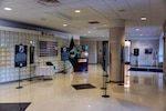 A display of posters with text white on black tells the stories of POW/MIA Day in a large vestibule area with doors to the outside on one side and a glass bottle wall on the other with an American Flag with the words We Are DLA above it. There is a table with a white cloth with a plate and other object on it. The table is empty. There is a vase with one rose as well on the table.