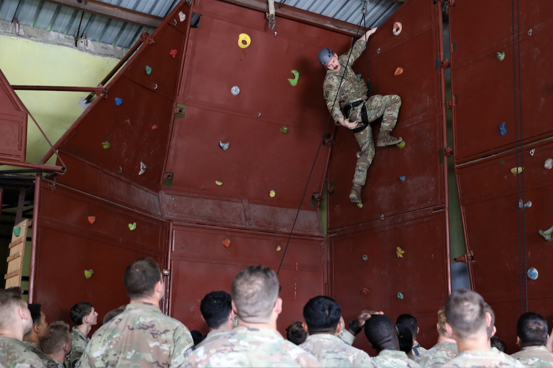 A soldier uses a rope to climb up a wall as others watch.