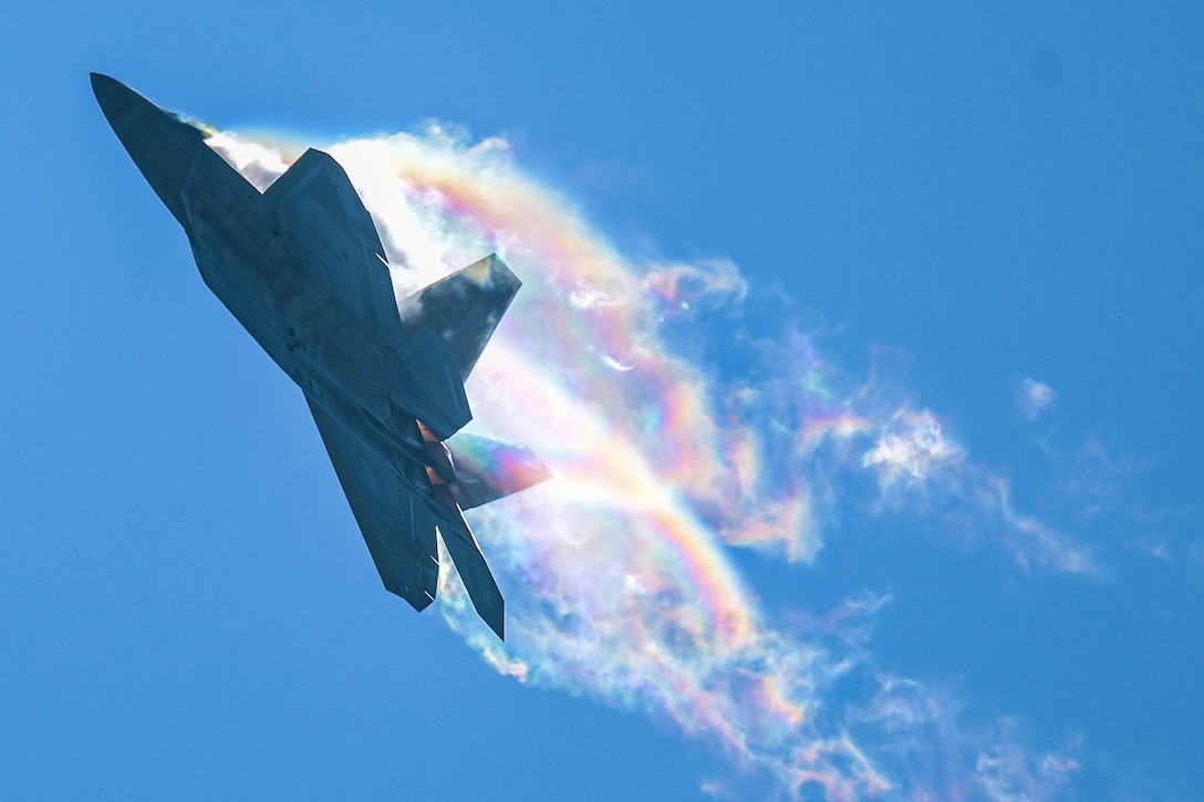 An Air Force aircraft leaves a vapor trail with rainbow highlights as it flies against a blue sky.