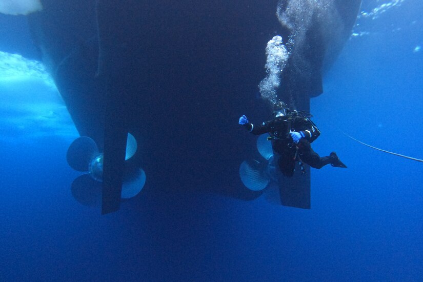 A military diver swims behind a ship.
