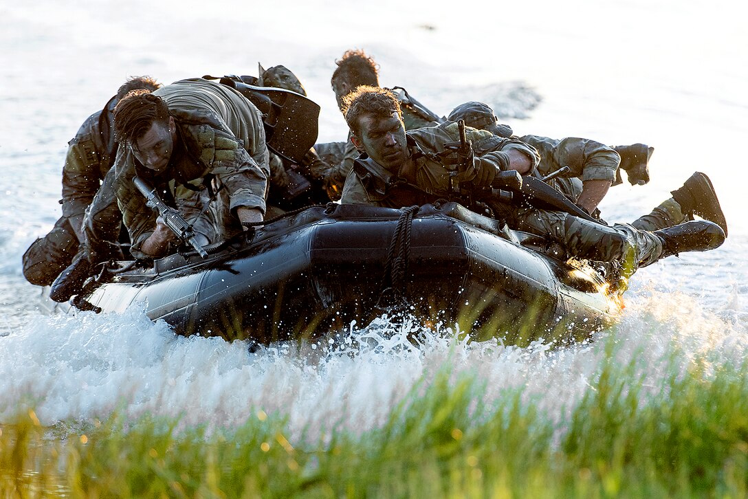 Airmen ride in a rubber boat that makes a splash as it approaches grassy shoreline.