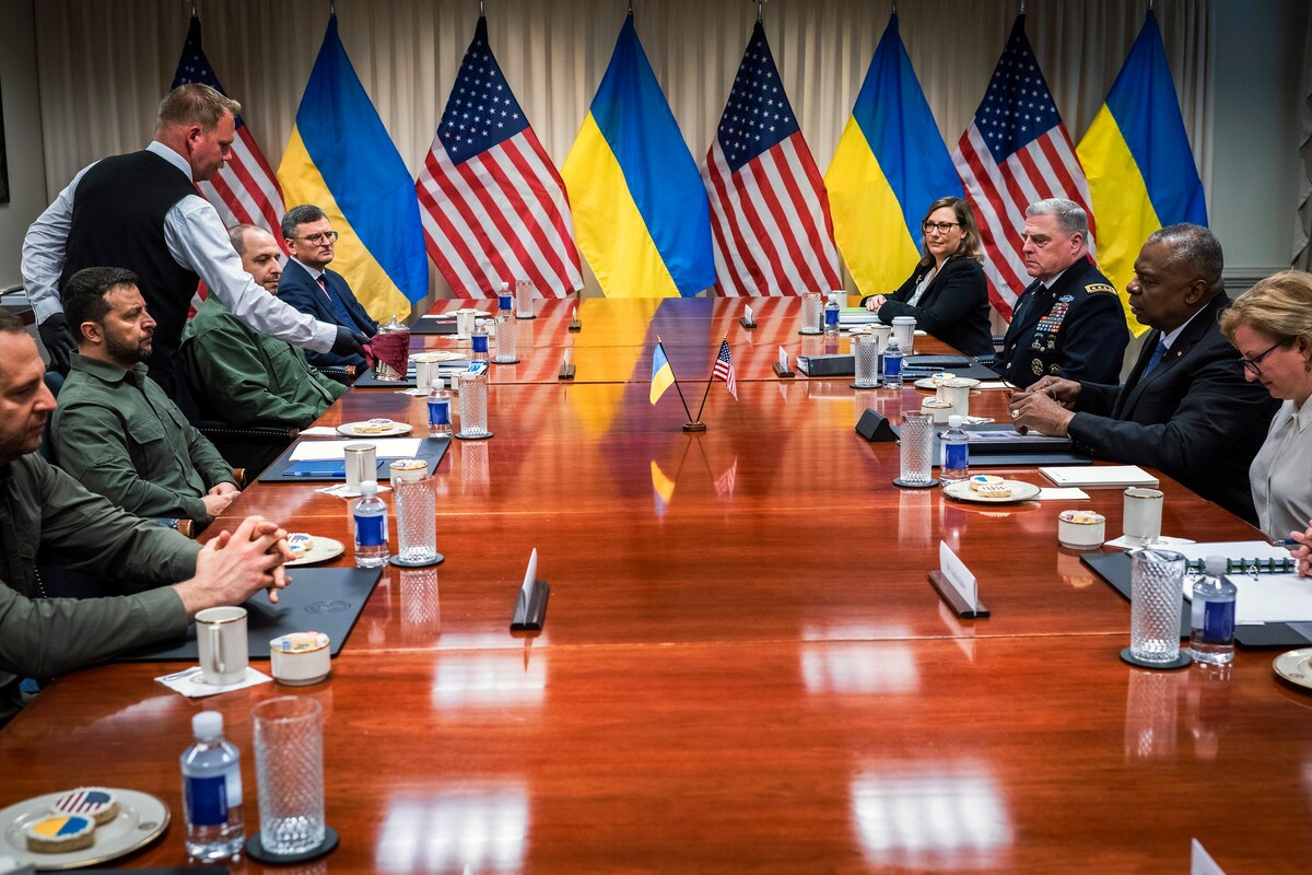 People in civilian attire and military uniforms sit around a conference table with U.S. and Ukrainian flags in the background.