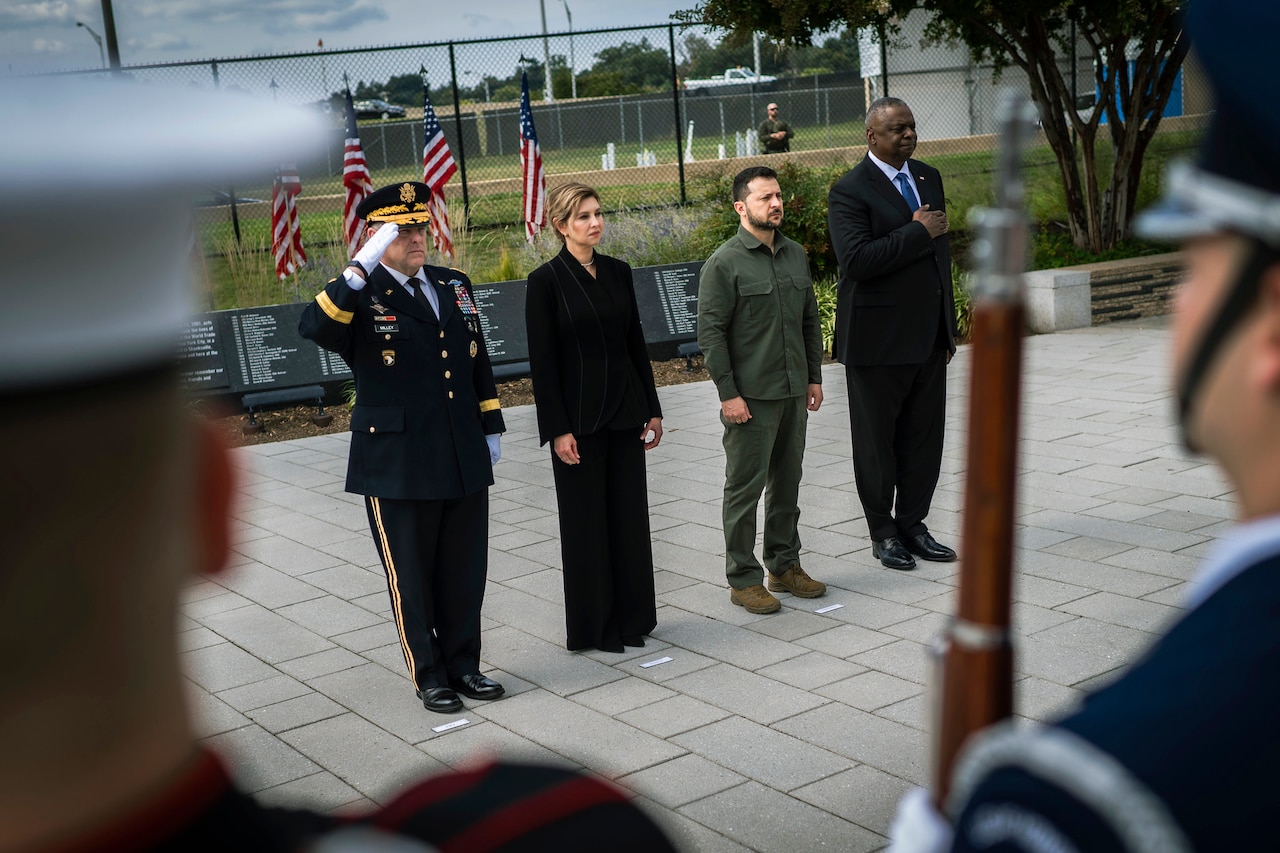 Four people stand in a row as face a military honor guard. American flags are raised behind them.