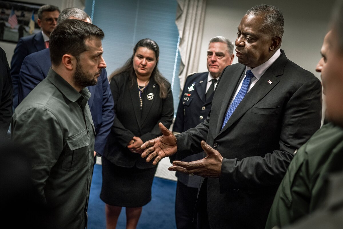 Two men speak, while others look on.
