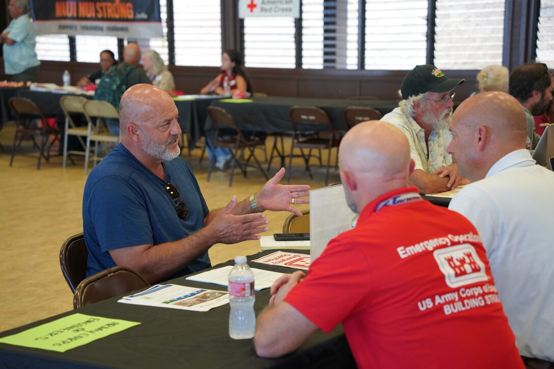 A man talks with two men who are seated at a table across from him