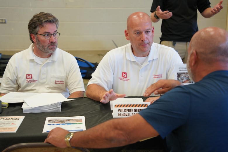 Two men wearing Army Corps of Engineers polo shirts talk to another man across a table from them.