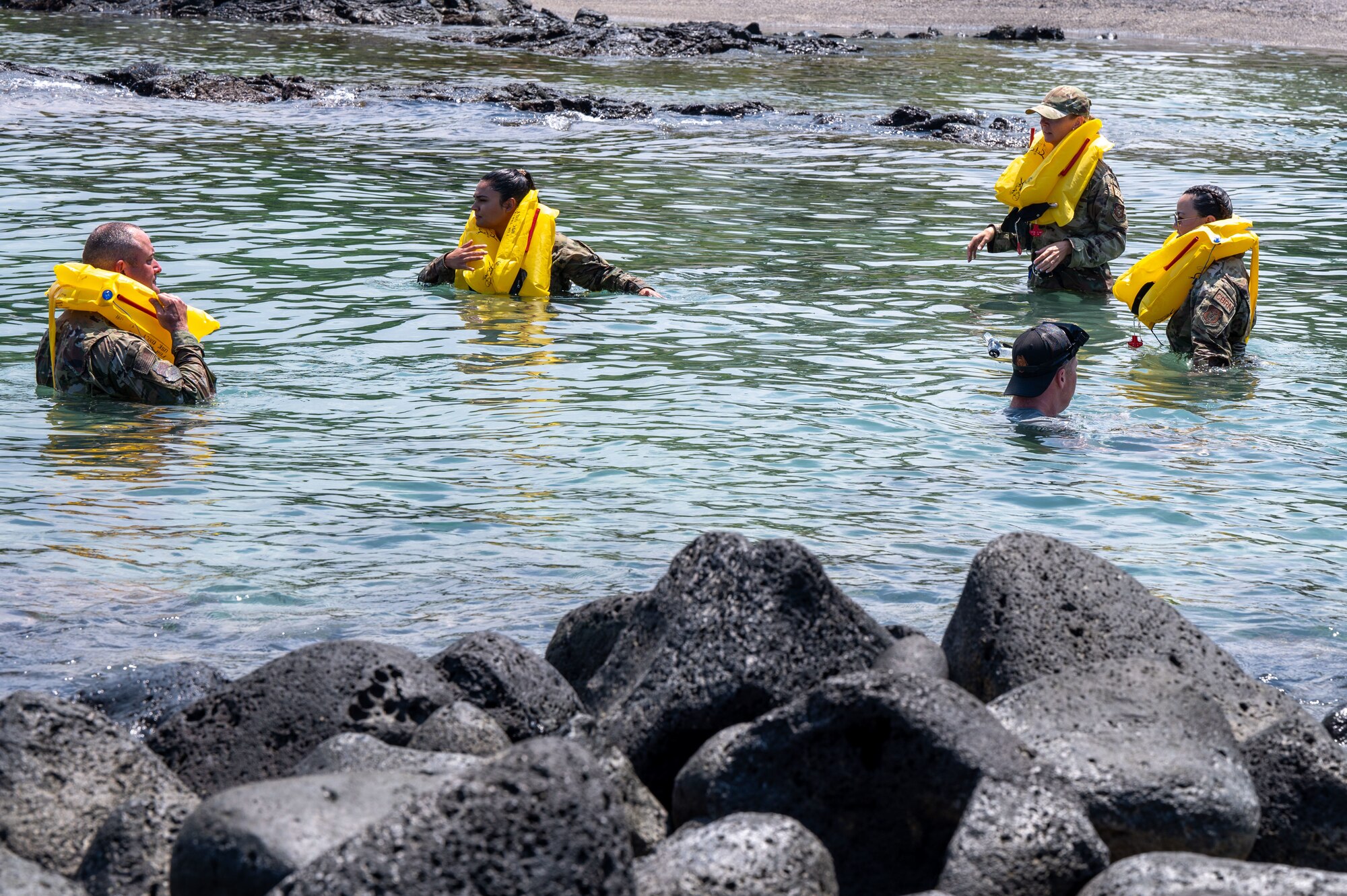 four people in military uniforms and yellow life vests stand in shallow water surrounded by rocks