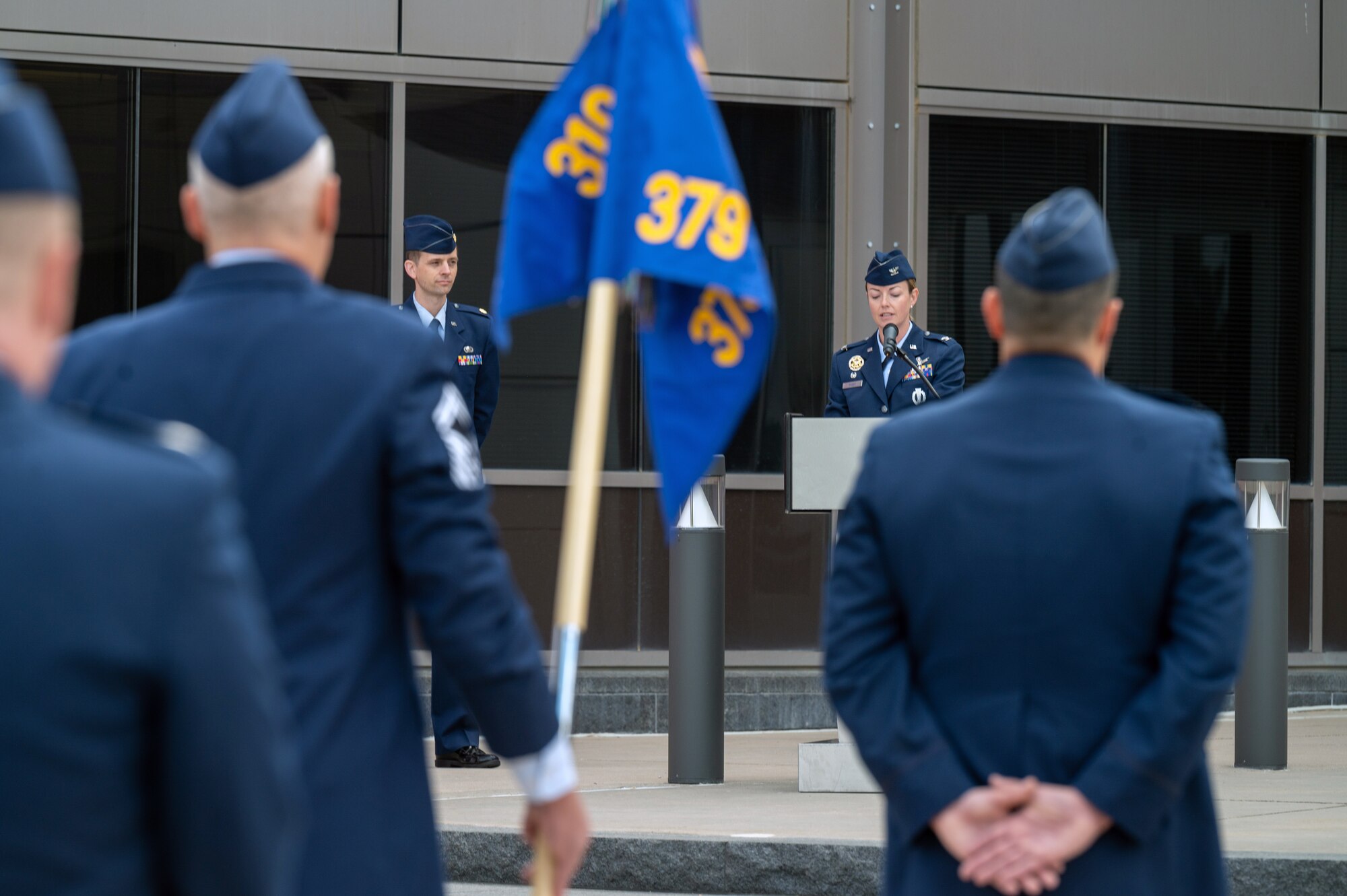 A group of people in military service dress standing outside a building holding a flag watch a woman in military service dress speaks into a microphone attached to a podium.