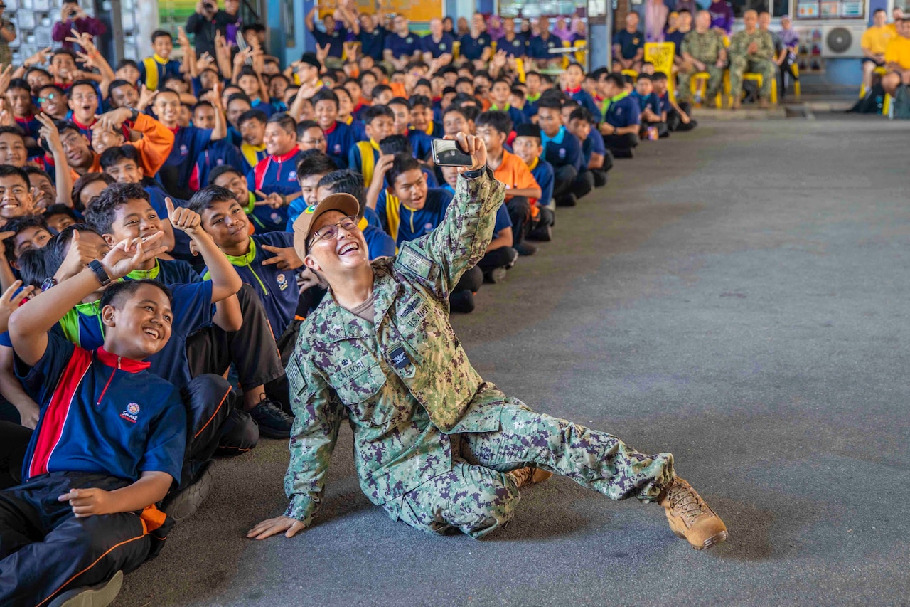 A sailors sits on the ground while taking a selfie with a large group of students.