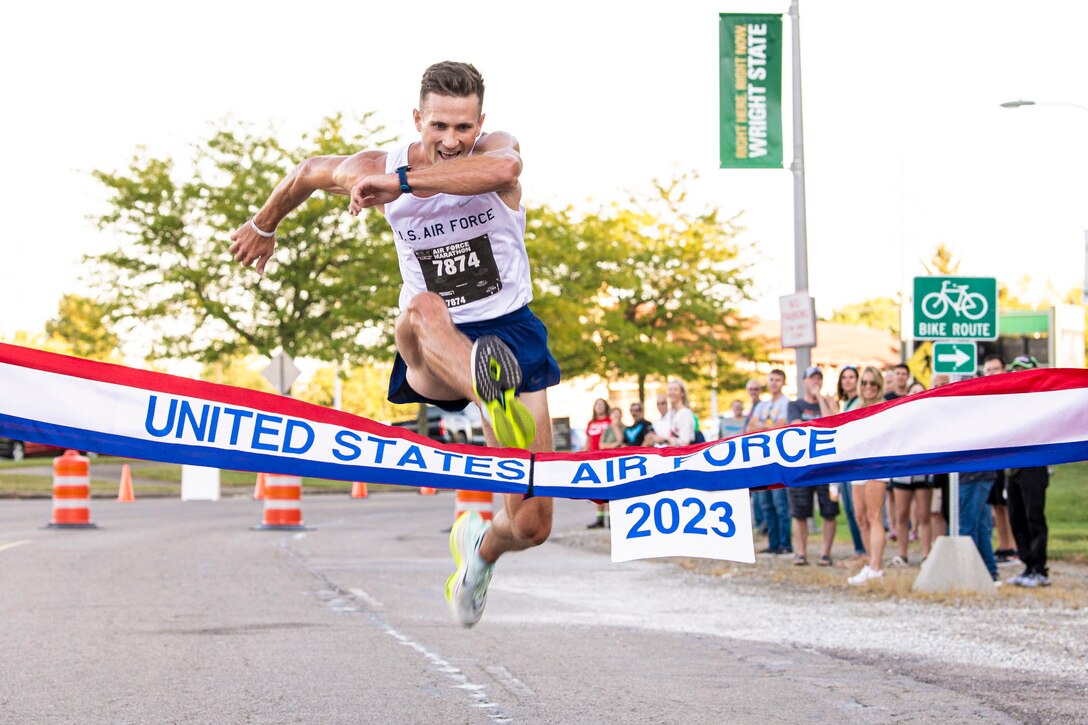 An airman jumps over a banner at the finish line of a race as spectators watch in the background.
