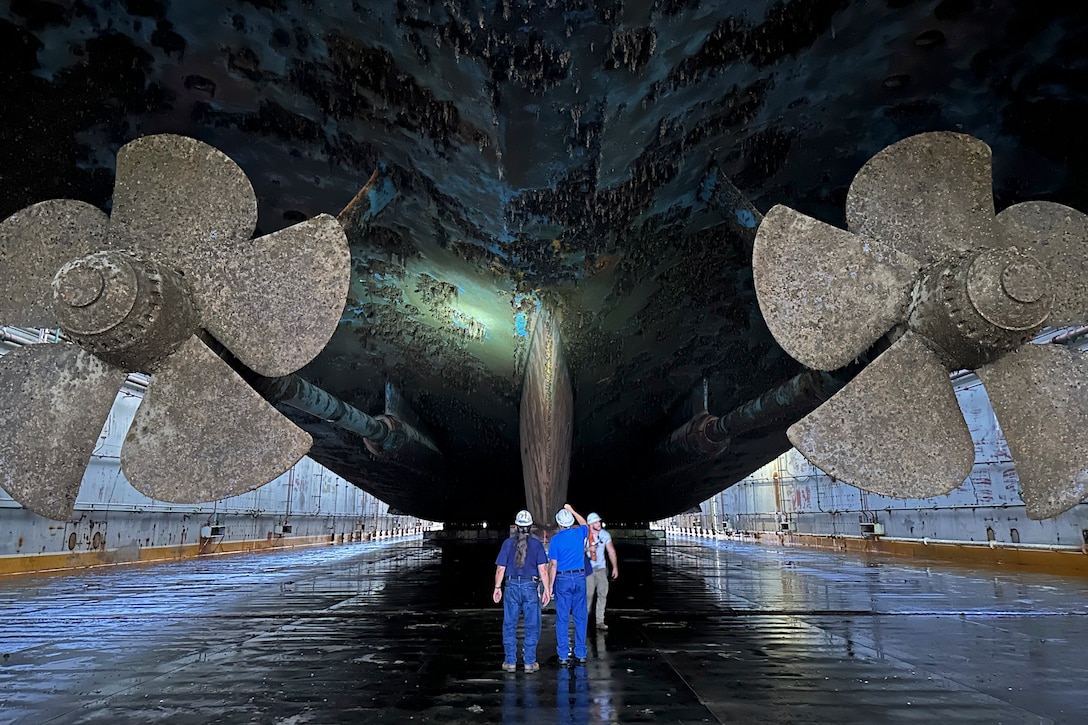 Workers wearing protective gear shine a light on a ship’s keel while standing in between two propellers.