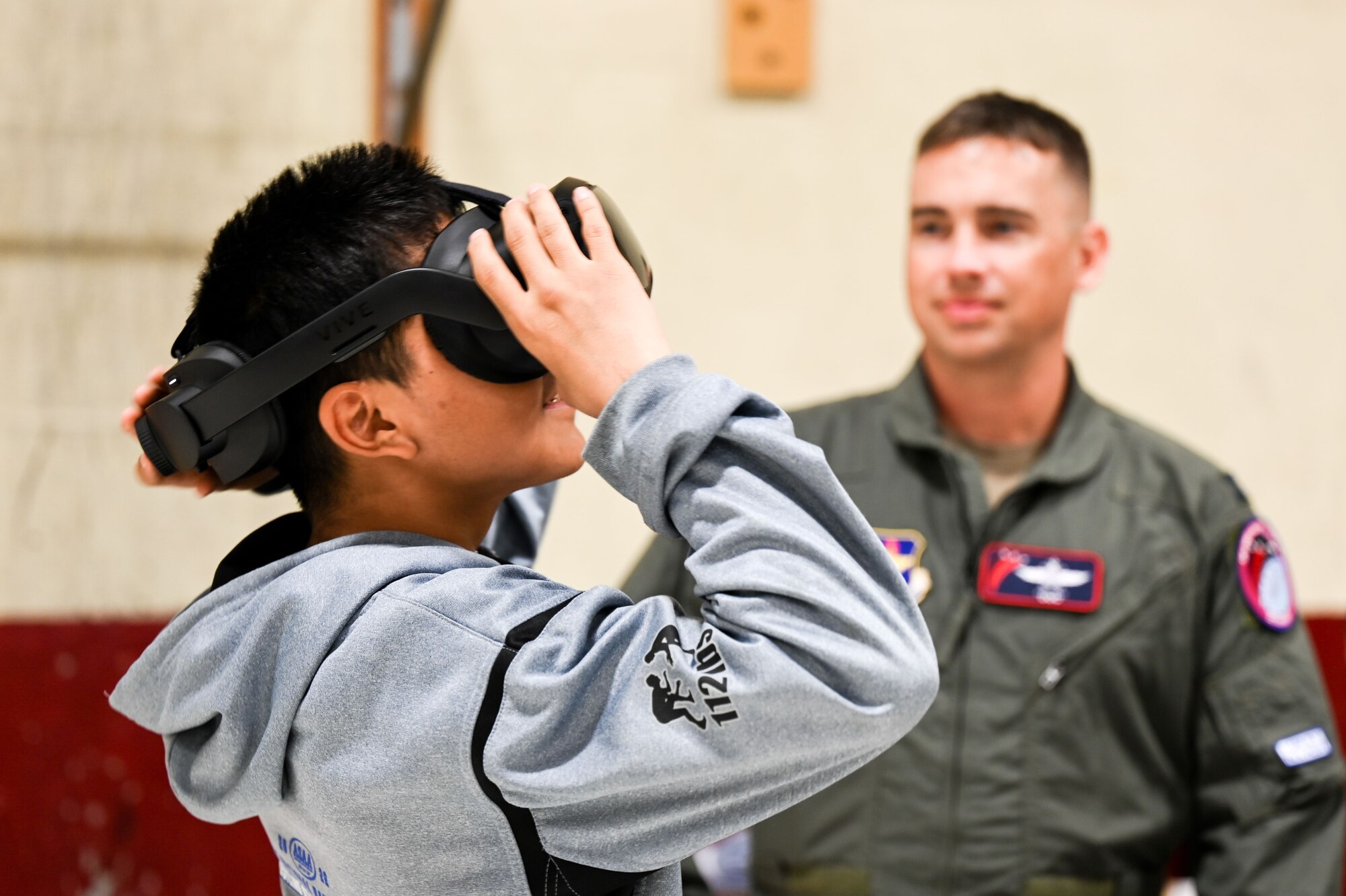 High school boy in grey sweatshirt looks through VR goggles while man in flight suite helps run the program