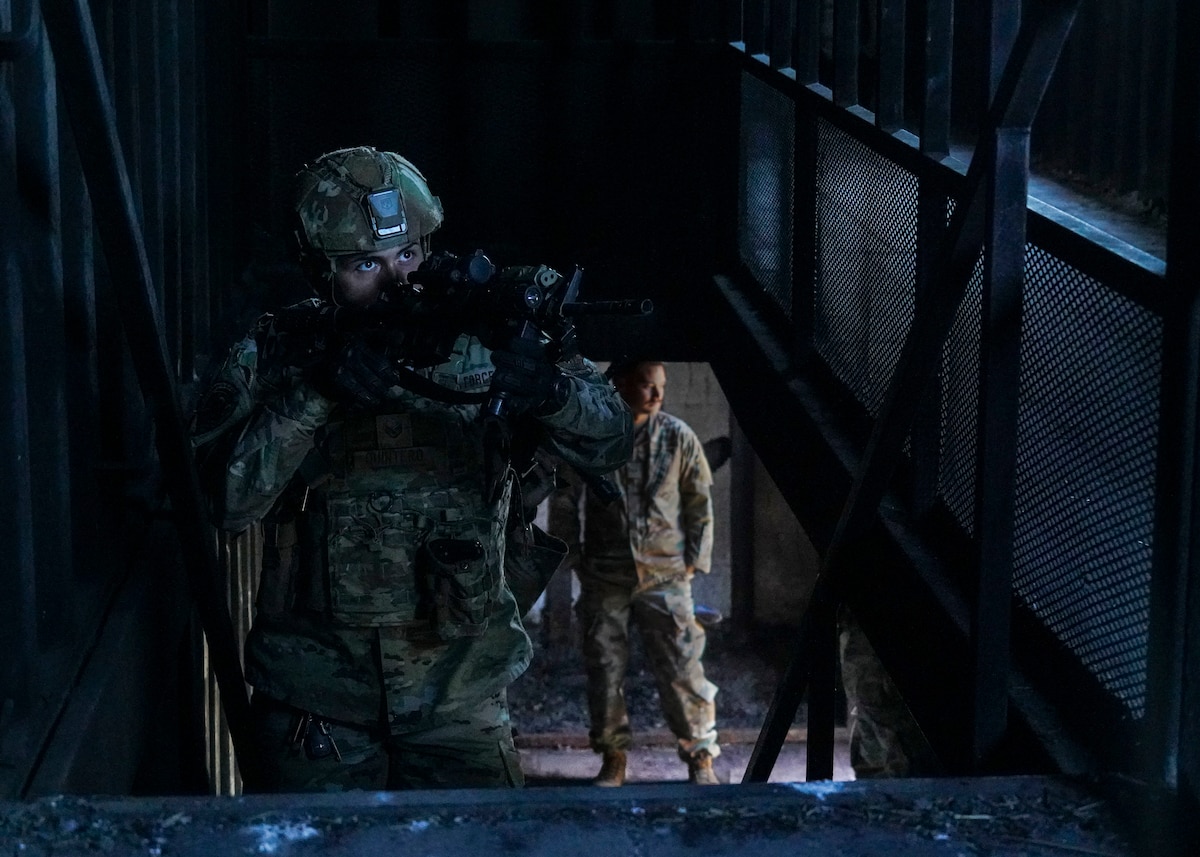A California Air National Guard Airman advances up a flight of stairs with his practice weapon drawn as he scans the room for threats in a dimly lit shipping container that has been retrofitted to serve as a building.