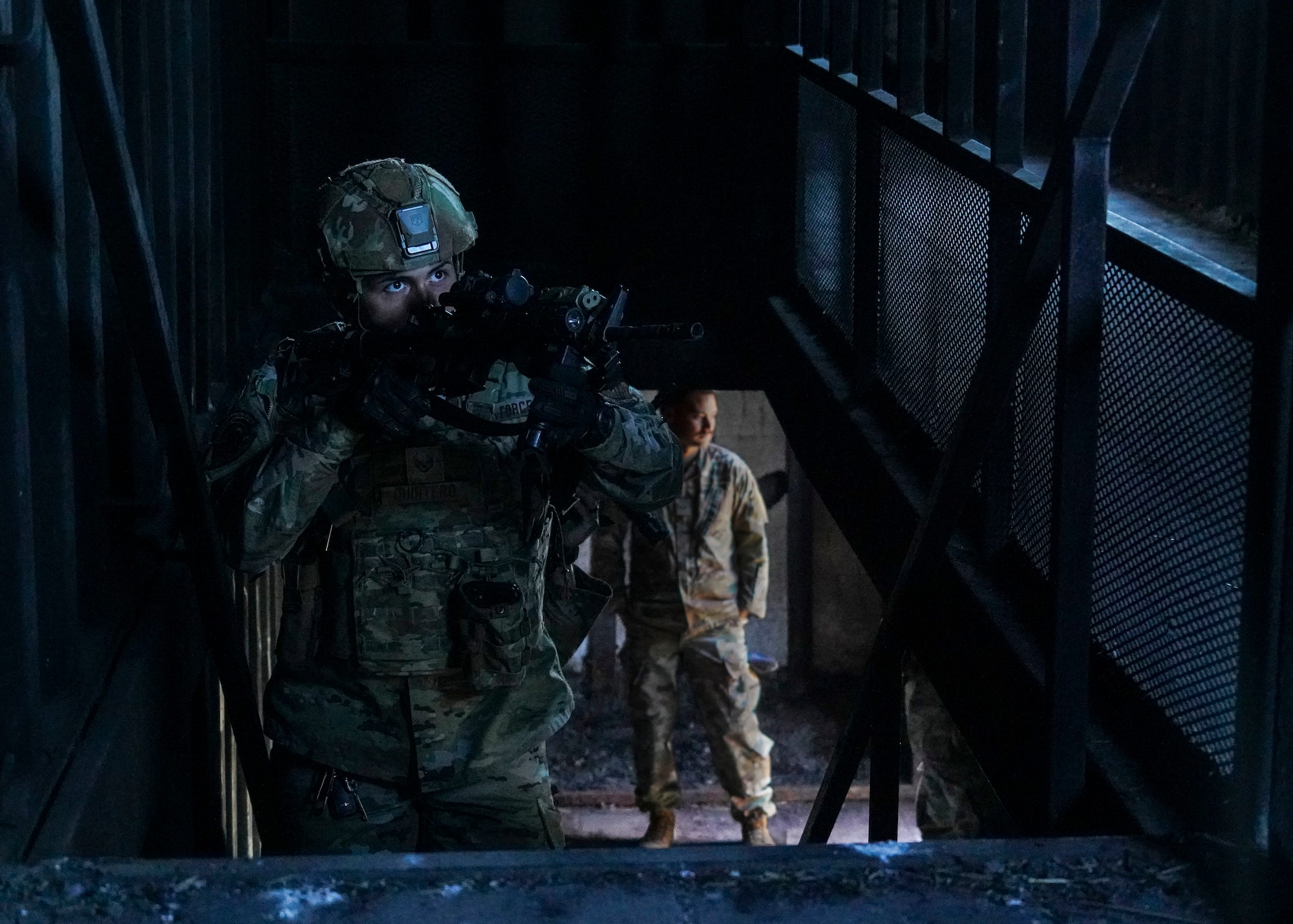 A California Air National Guard Airman advances up a flight of stairs with his practice weapon drawn as he scans the room for threats in a dimly lit shipping container that has been retrofitted to serve as a building.