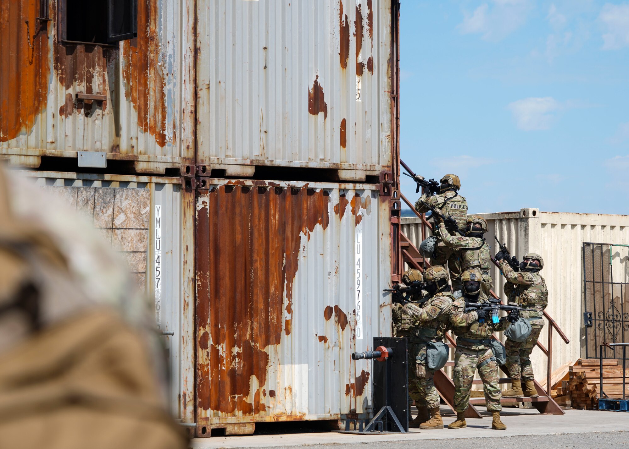 A team of six California Air National Guard Airmen move up a flight of stairs attached to a shipping container that has been retrofitted to act as a building. The Airmen in the photo are in the distance towards the left side of the image.