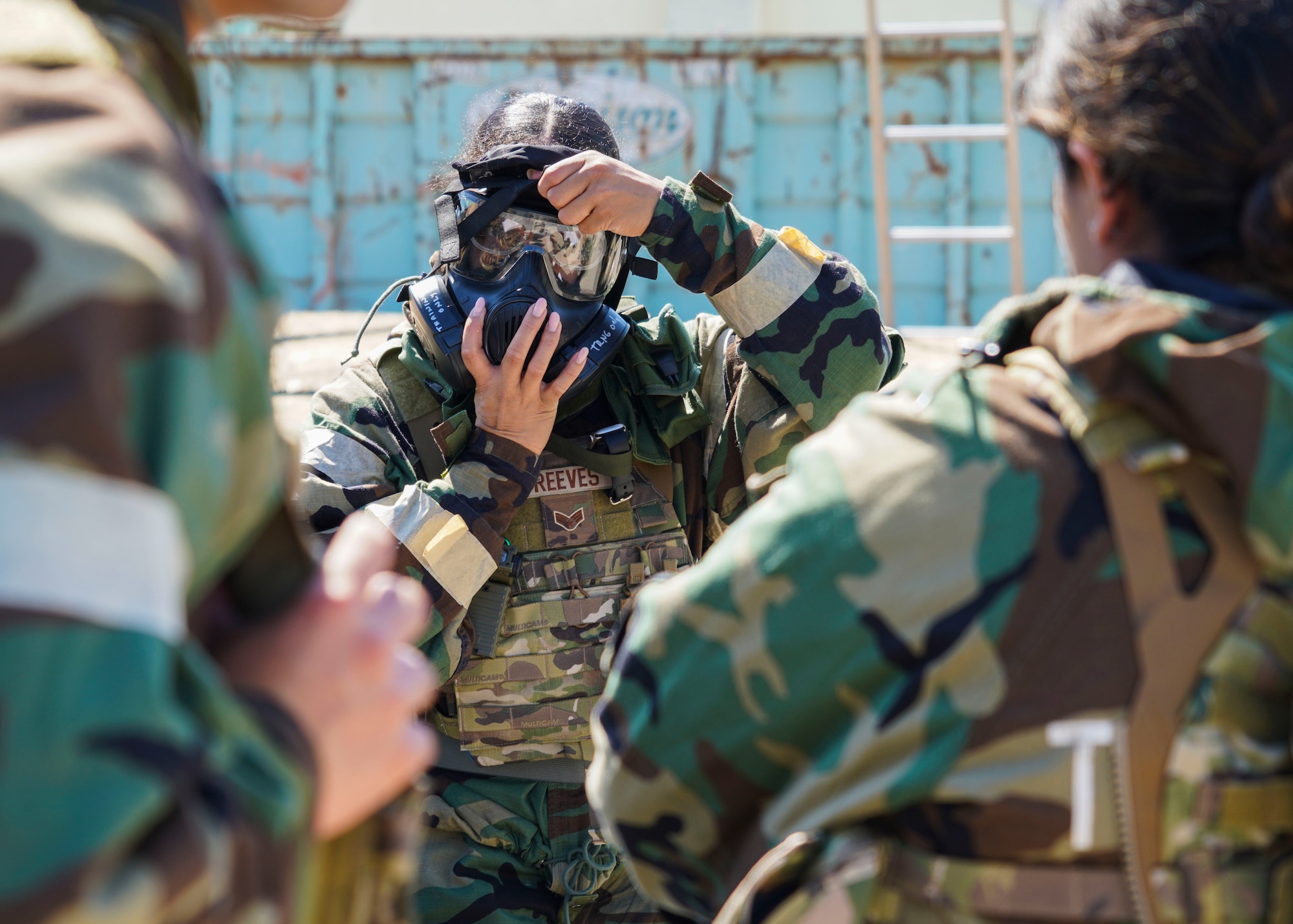 A California Air National Guard Female Airman is framed compositionally between two other Airmen in chemical protection gear as she dons on her chemical gas.