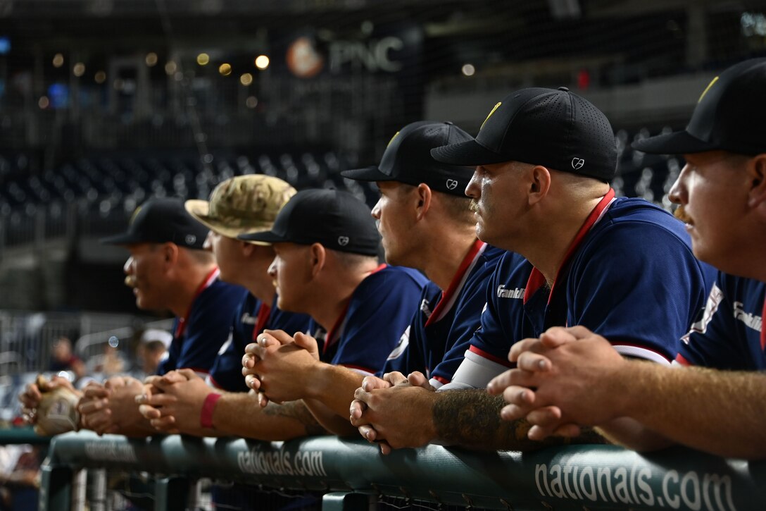 Six players of the Joint Base Andrews softball team lean on a railing while watching their teammates play from the sidelines.