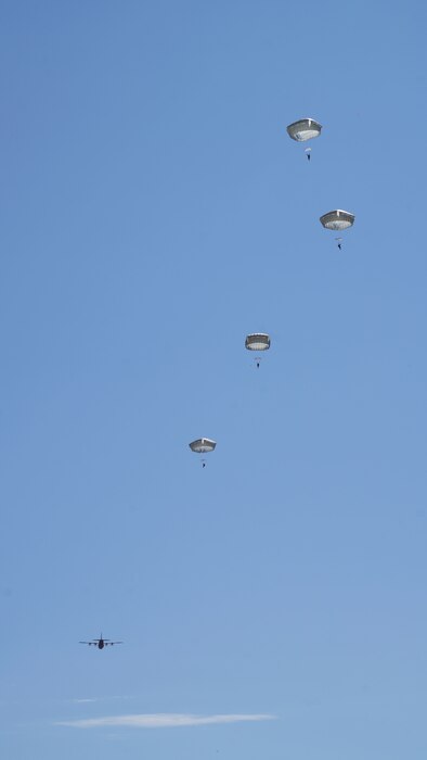 A photo of multiple U.S. Army paratroopers who have deployed their parachute after jumping from a C-130J pictured off in the distance affixed against a blue sky.