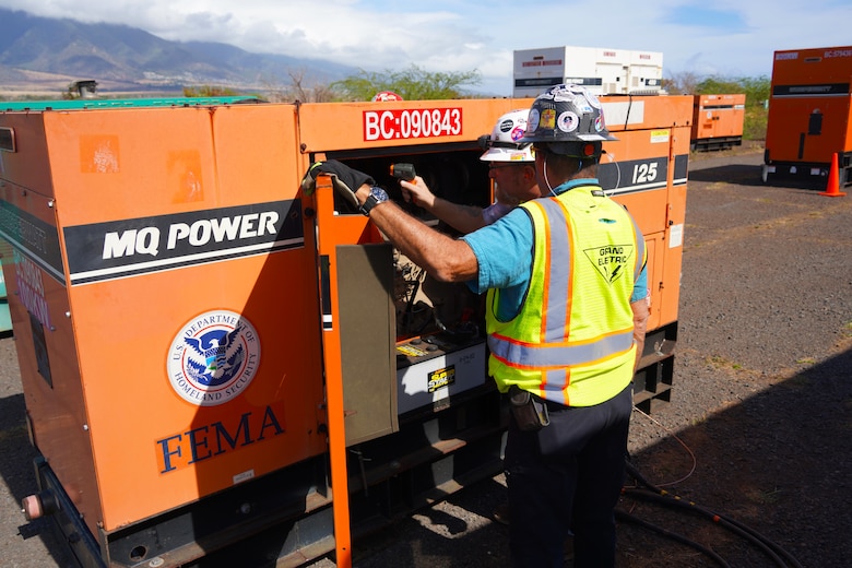 Two men in safety gear inspect an industrial power generator with the FEMA logo on it