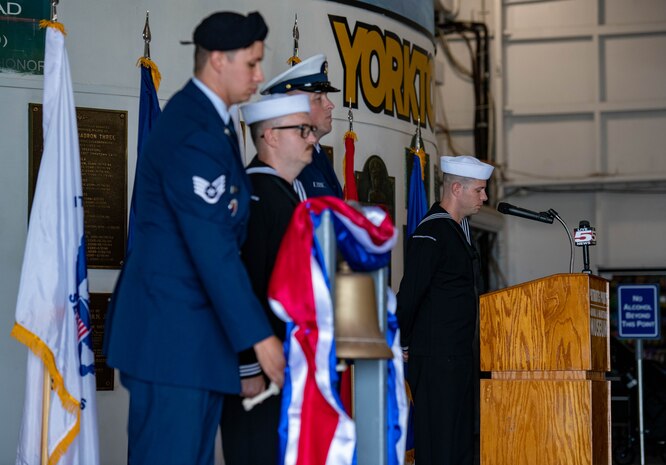 A U.S. Navy Sailor reads the names of fallen service members as a U.S Air Force Airman rings a bell, during a Bells Across America ceremony aboard the decommissioned aircraft carrier USS Yorktown