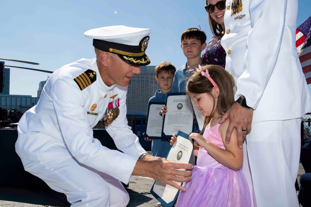 A sailor hands a child a paper as another sailor stands above holding her shoulders next to a woman and two children.