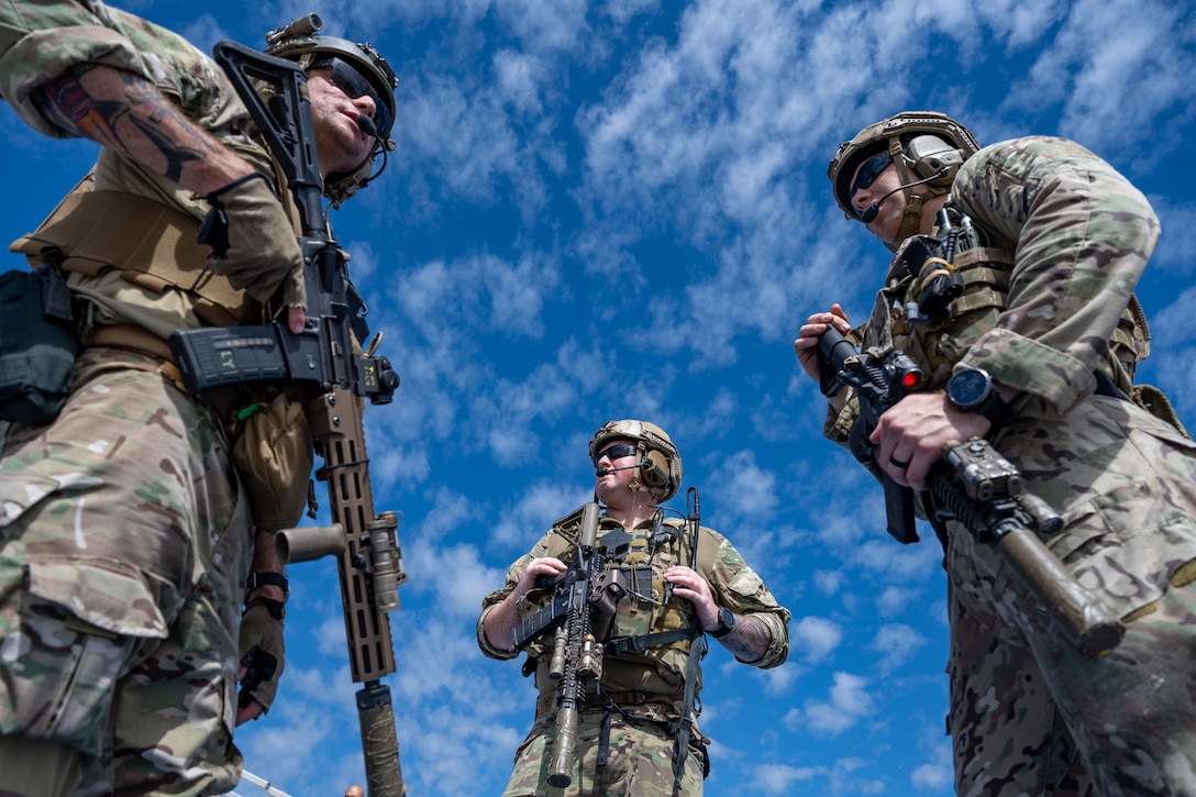 Three uniformed airmen are photographed from below while standing in a semicircle holding their weapons.