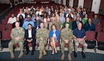 A large group of smiling people sit in an auditorium facing the camera. Some are in business attire and some are in military uniforms. There are men and women.