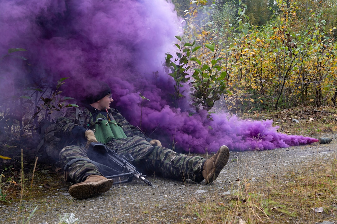 A cadet lays in a field while carrying a weapon as a flare releases purple smoke to the right.