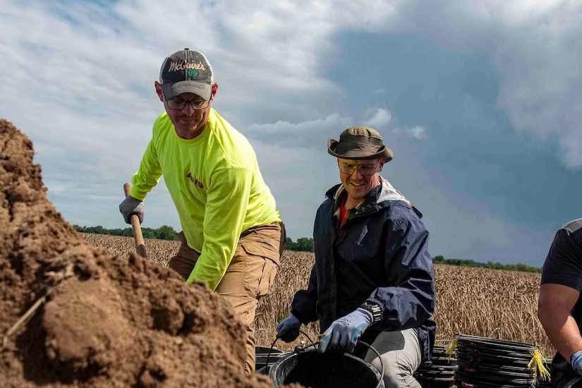 Men shovel dirt, then remove it in buckets. Behind them is a field.