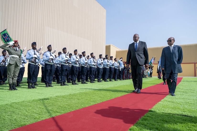 Two men wearing suits walk along a carpet as members of a foreign military stand in attention.