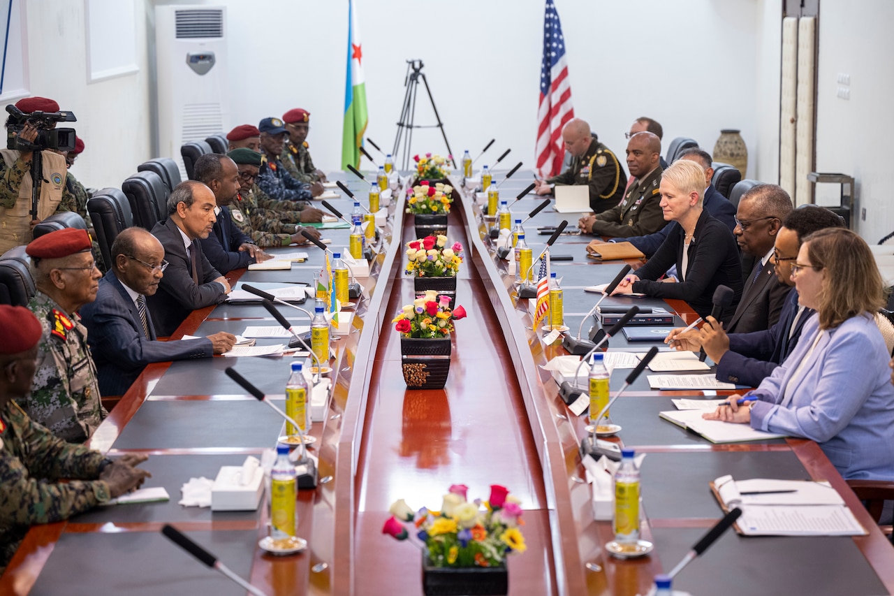 Men and women wearing civilian attire and military uniforms sit around a long conference table.