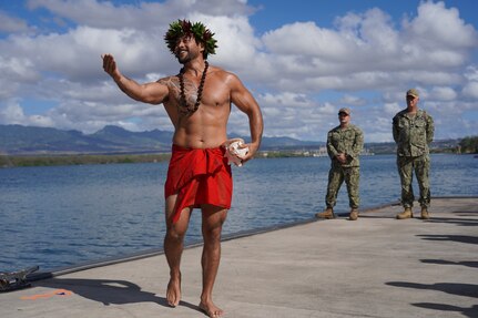 Louis Santos, extends his right arm to the left of the frame while holding a conch shell on his left hip, as he performs a traditional Hawaiian oli (chant) to bless the land, asking for protection and to foster good energy, during a Hawaiian blessing ceremony held at the water front of Pearl Harbor, as ceremony attendees look on.