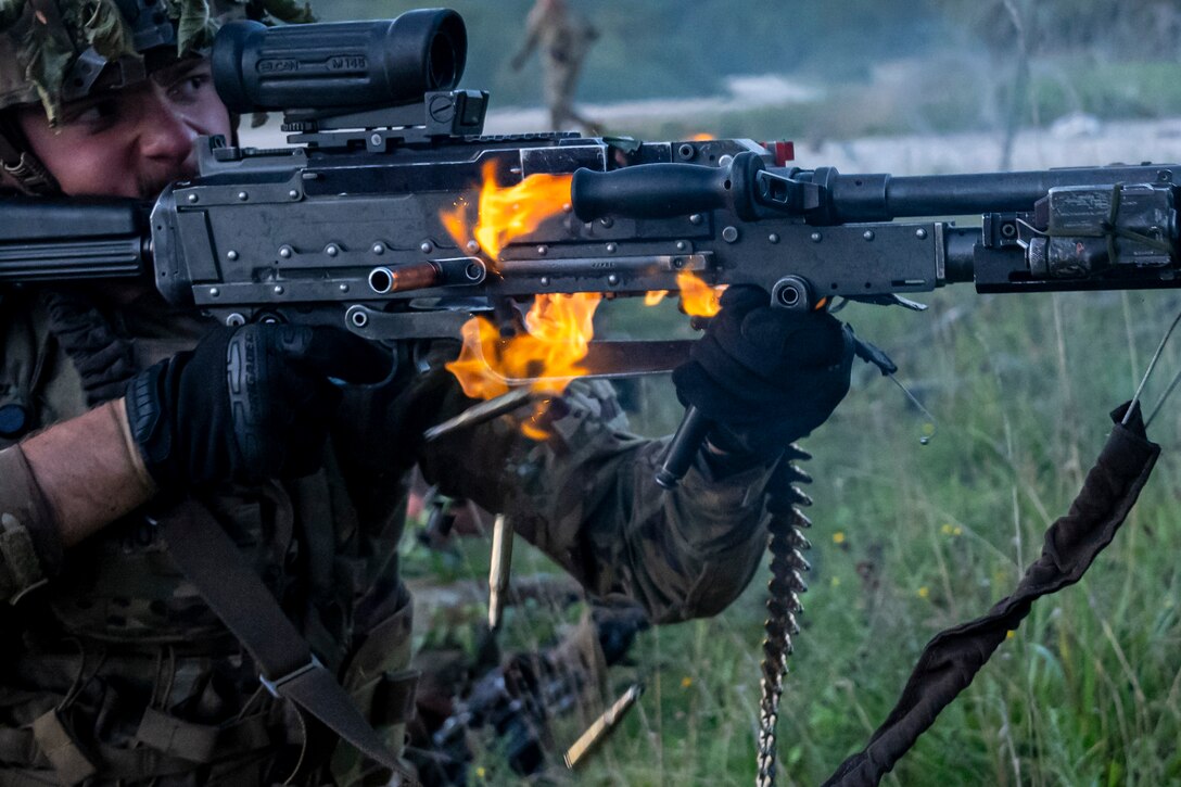 A soldier looks through the scope of a weapon with flames coming out of the side of it while firing it.