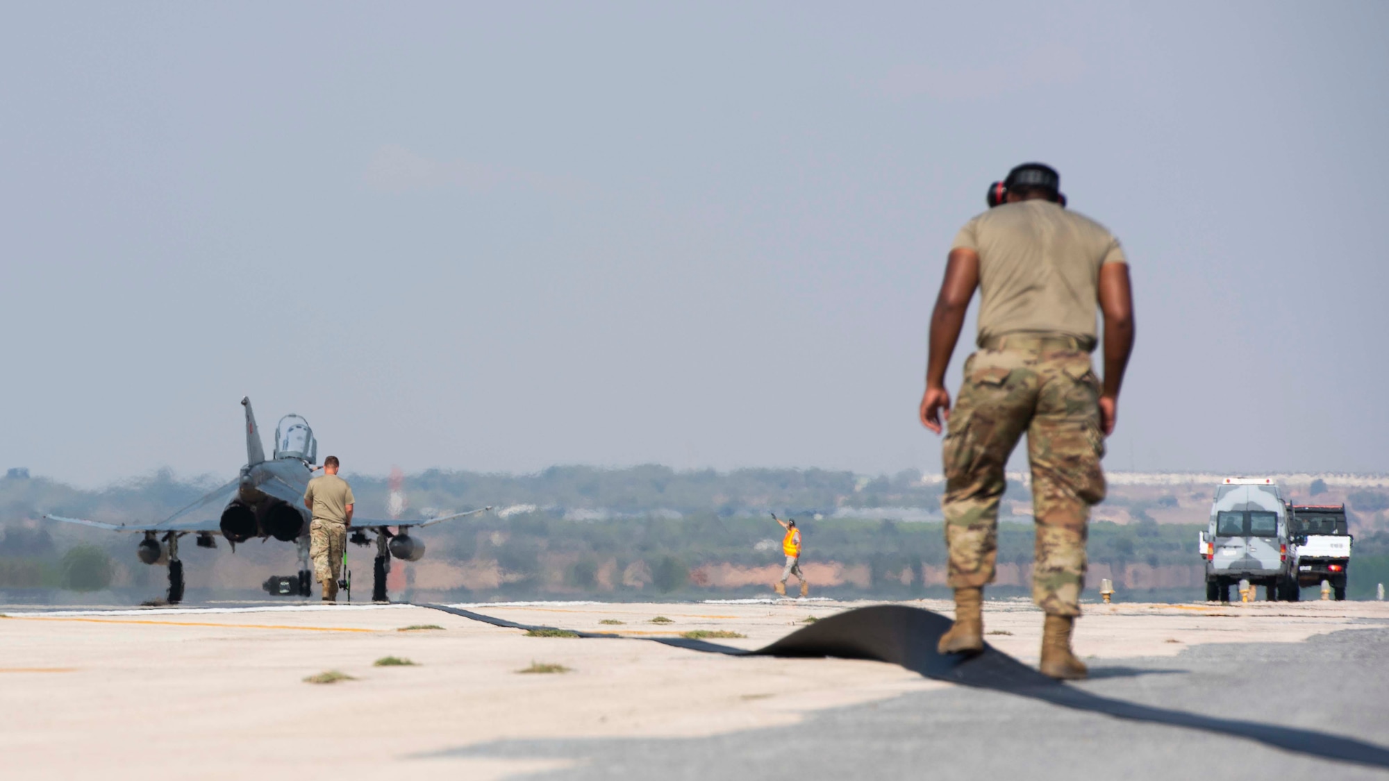 A fighter jet and two men can be seen next to a large cable stretching across a runway