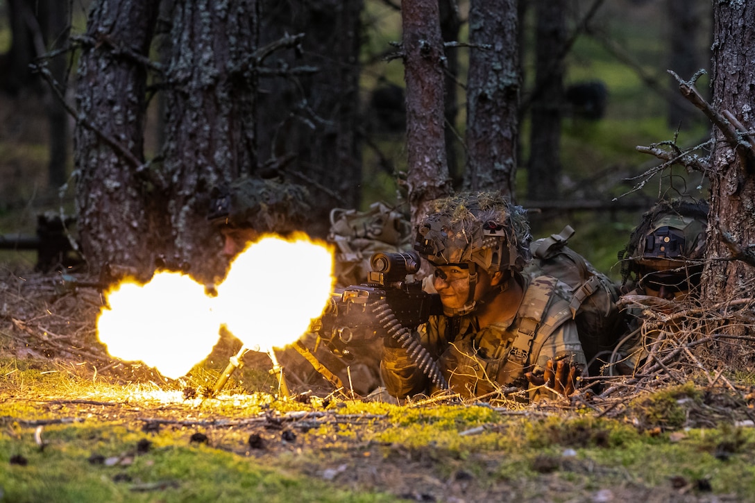 A soldier lays on the ground looking through the scope and firing the weapon while fire blazes out of the barrel.