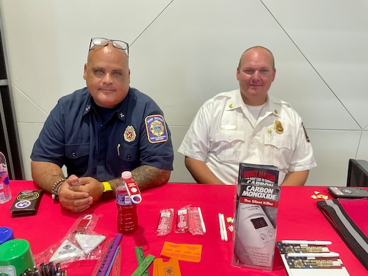 Two firefighters sit at a table that has information about fire safety.