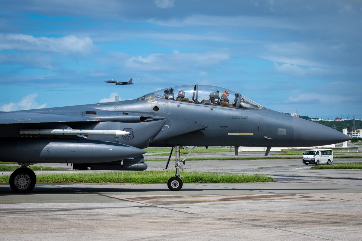 A U.S. Air Force F-15E Strike Eagle assigned to the 391st Fighter Squadron, Mountain Home Air Force Base, Idaho, taxis as another F-15E assigned to the 336th Fighter Squadron, Seymour Johnson AFB, N.C., soars over Kadena Air Base, Japan during a no-notice agile combat employment exercise, Sept. 22, 2023.