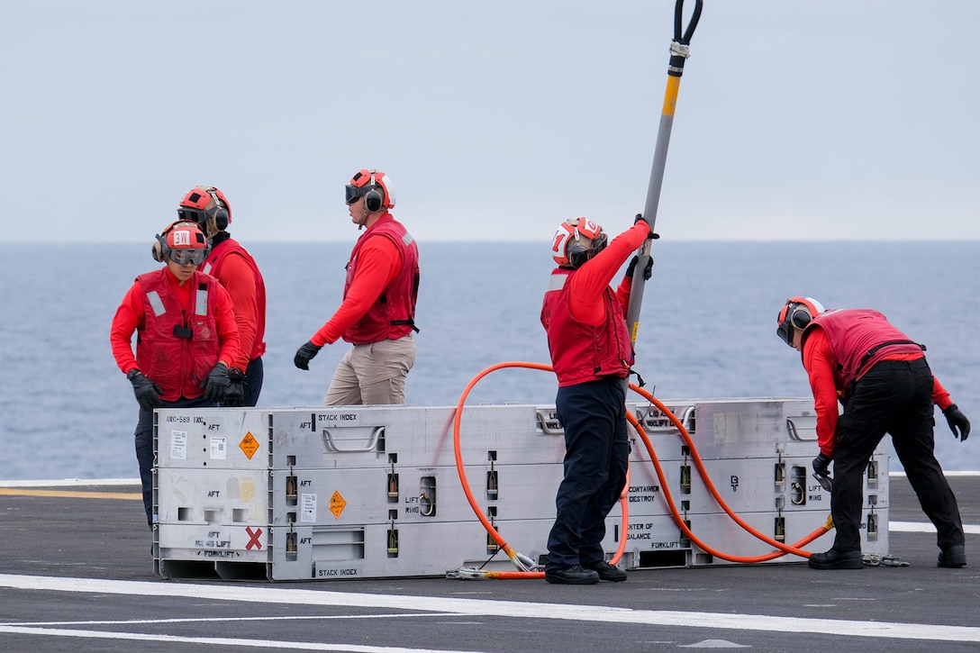 Two sailors remove stabilizing lines from one side of an ammunition crate and three others stand behind it.