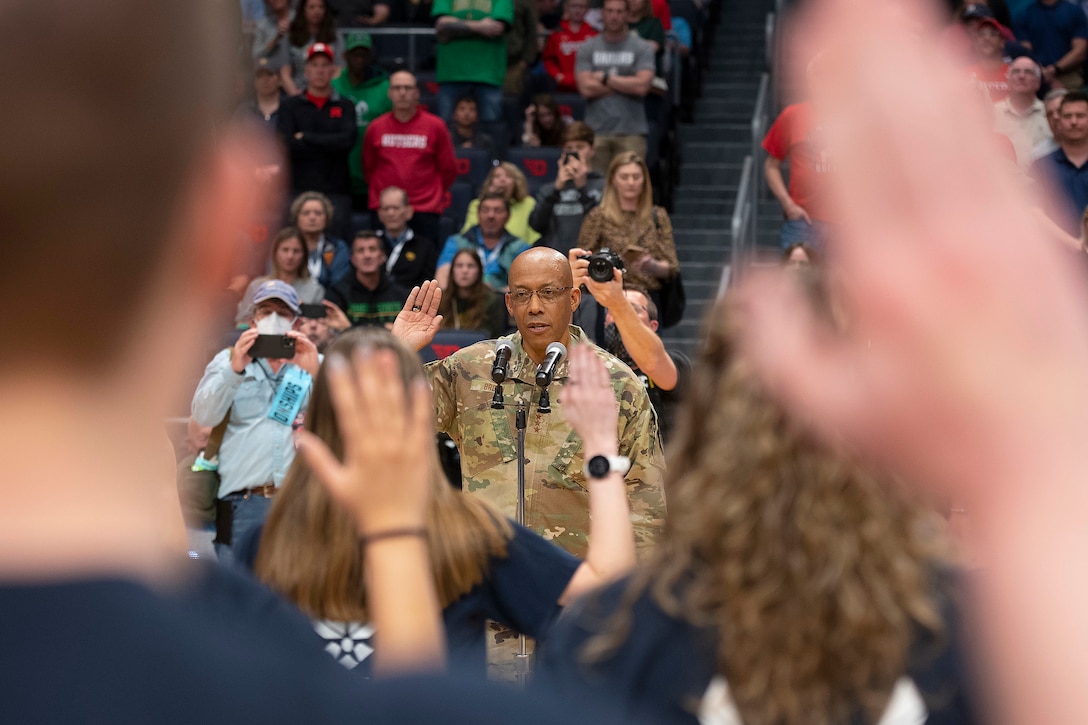 An Air Force general stands with right hand raised facing a group of recruits doing the same in front of a crowd.