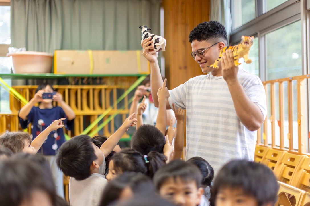 A Marine smiles while holding two animal figurines in the air as a dozen preschool children look or point at them.