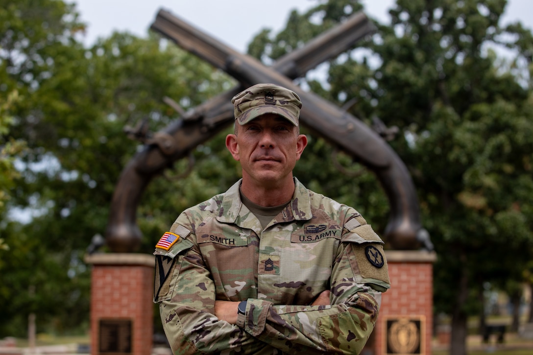 An Army Soldier poses for a photo in front of crossed rifles in the background, a symbol of the Army's Military Police.