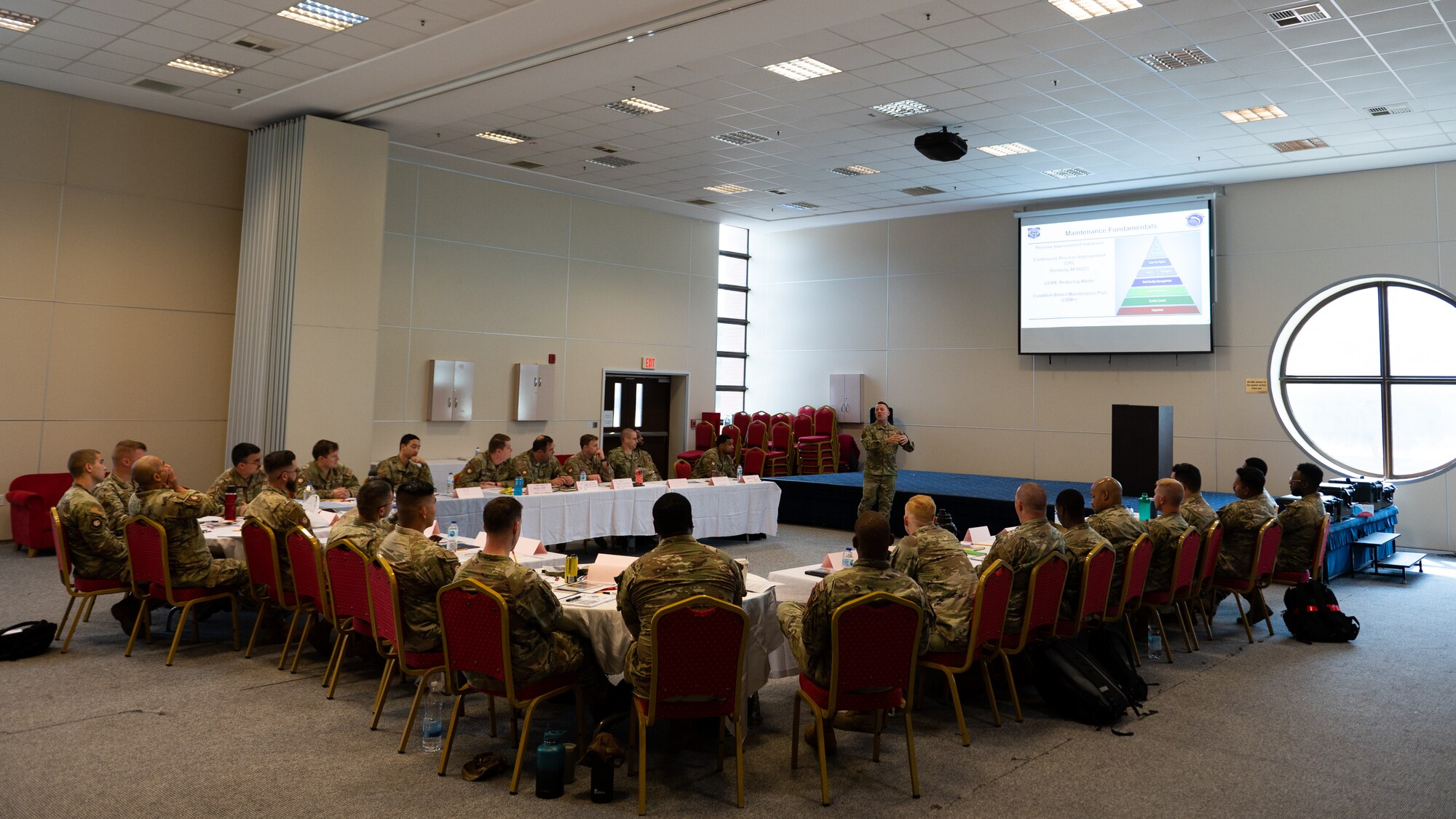 An instructor addresses a large room of Airmen seated in a  square around him