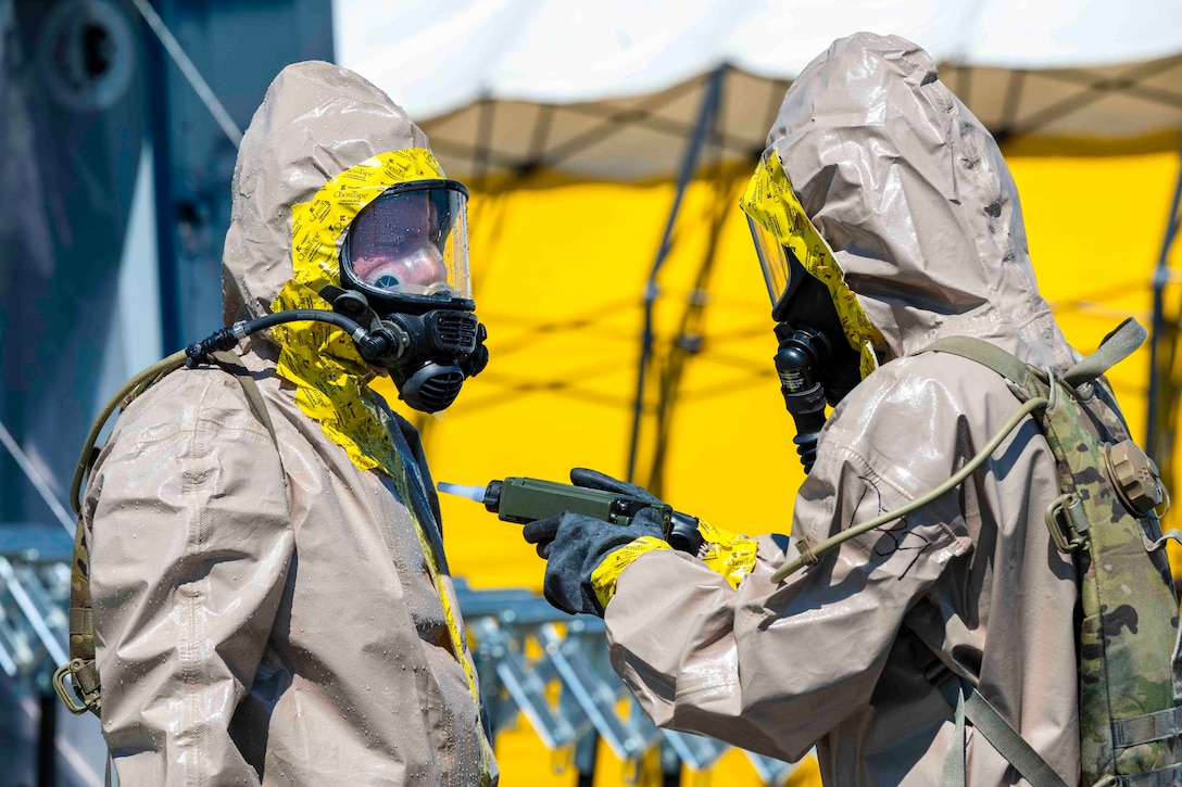 Two guardsmen wearing hazmat suits face each other as one uses a piece of equipment.